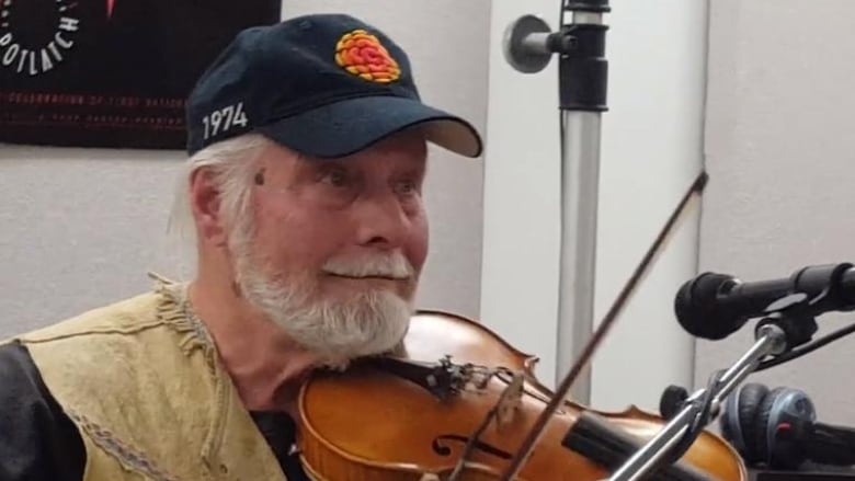 A bearded man in a CBC hat holds a fiddle.