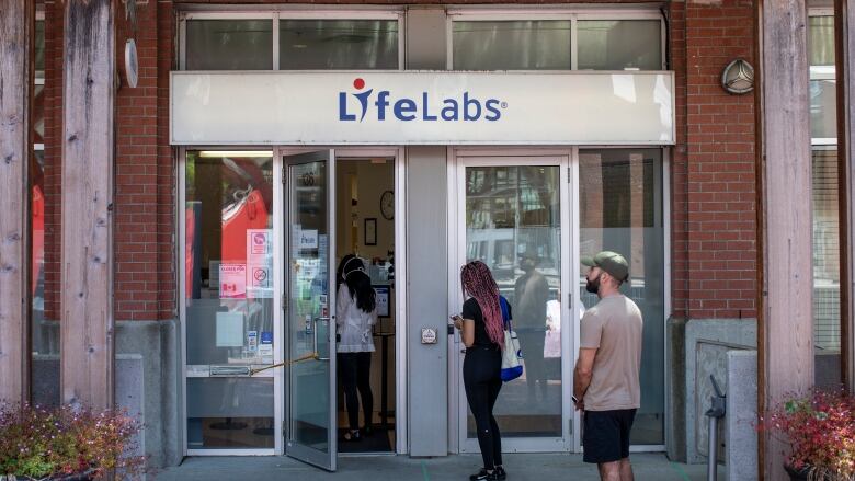 People wait in a lineup to enter a LifeLabs location in a brick building