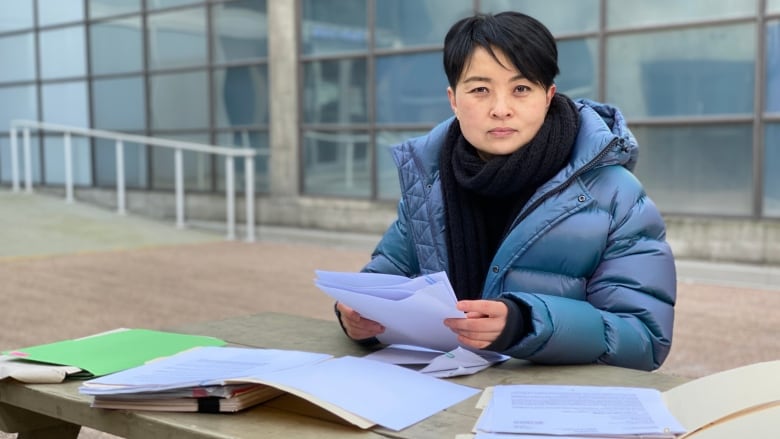 A woman sits at a bench outside, papers spread in front of her.