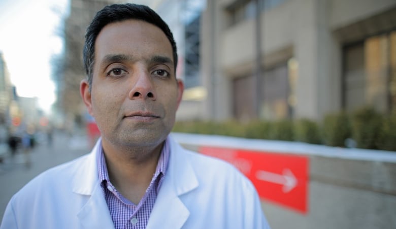 Dr. Samir Sinha stands outside a hospital, wearing a white lab coat. He's looking directly into the camera.