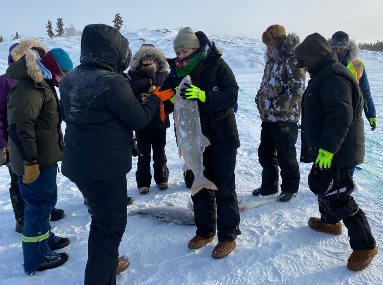 A group of people stand around a hole in the ice. One of them holds a large fish.