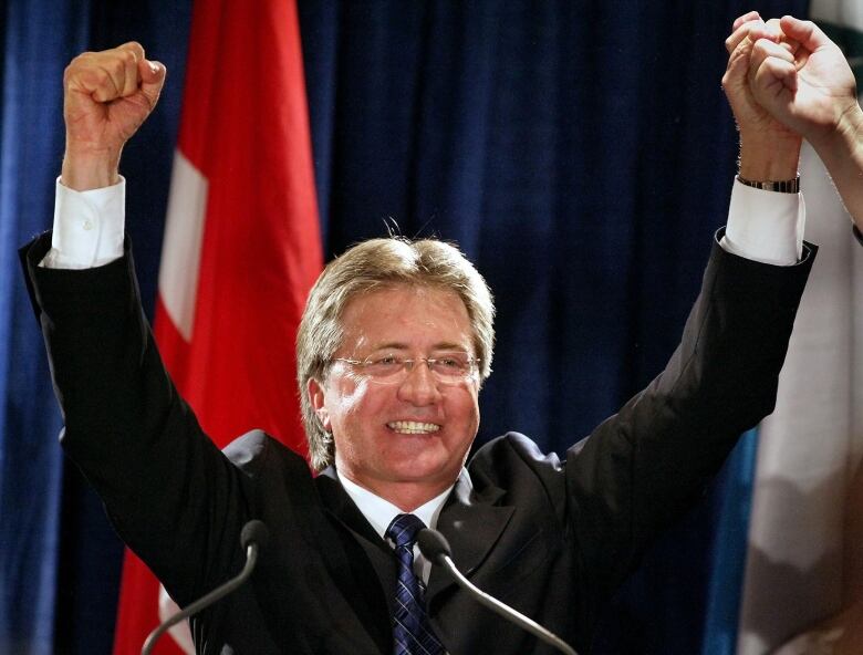 A smiling man with grey hair wearing a suit, with his hands in the air.