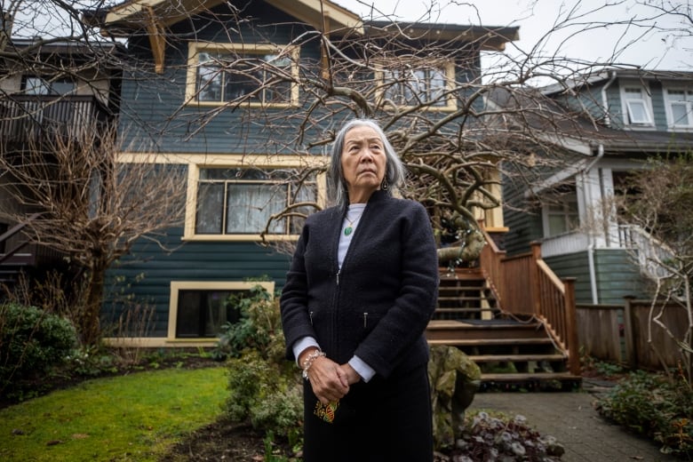 An Asian woman stands outside a heritage home with a tree in the yard, looking serious.