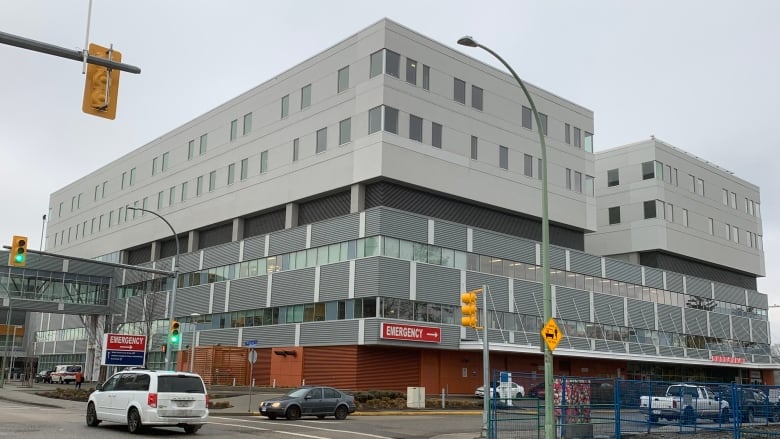 A large four-storey square building with a red Emergency sign on the front corner faces an urban intersection. 