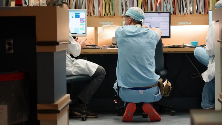 A physician in full hospital scrubs looks over his shoulder at a coworker.