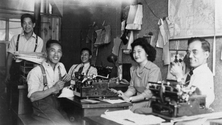 A black and white photo of five people in a newsroom