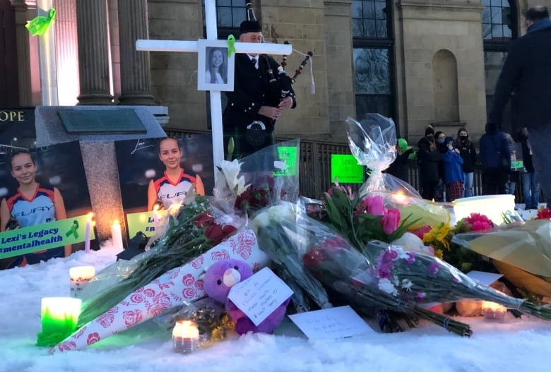 Flowers, candles and pictures lay in the snow as a bag pipe player stands in the background in front of an official looking building.