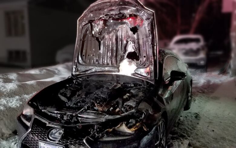 A car parked outdoors at night with snow in the background has its hood up, revealing fire has burned the engine area.