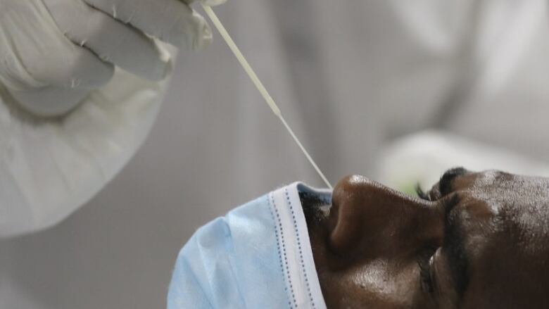 A health-care worker wearing a white medical gown and white gloves collects a nose swab sample for a polymerase chain reaction (PCR) test from a man whose blue medical mask is pulled down to cover his mouth only.