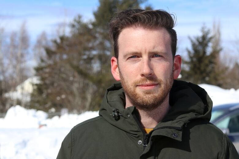 A man wears a winter jacket outside with snow in the background.