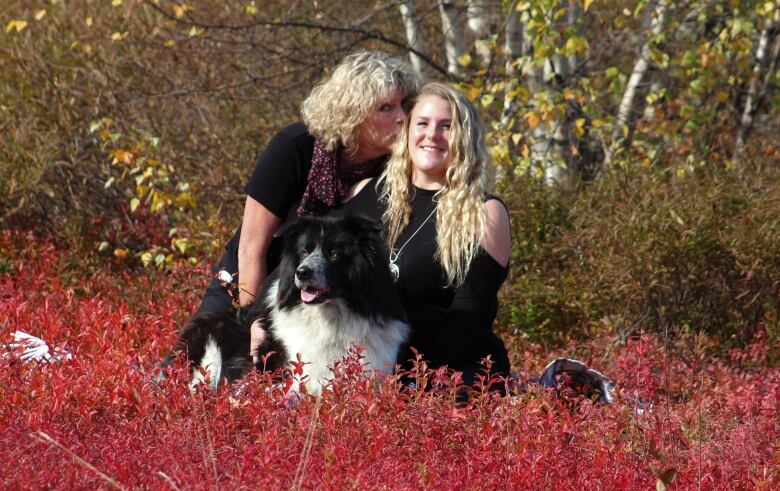 A mom snuggling her daughter in the midst of a brilliant-red field of blueberry leaves while the daughter hugs a large, black and white dog. 