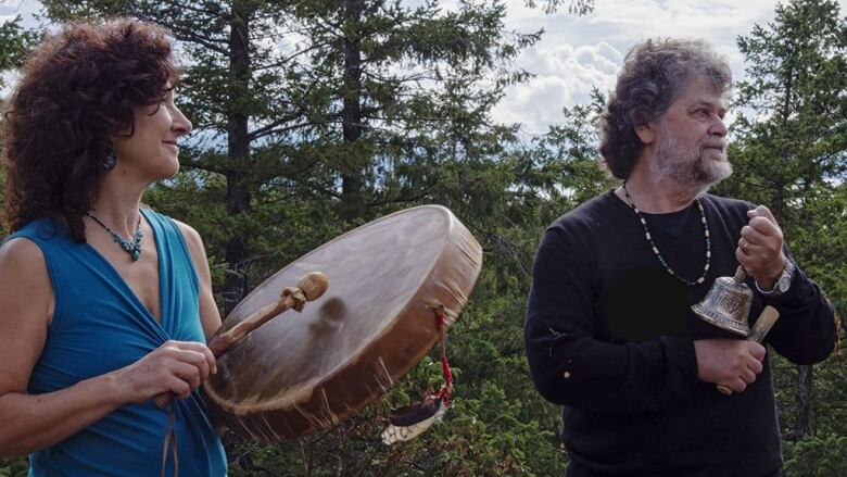 A white woman pounds a drum next to a white man holding a bell. They are standing in a forest.