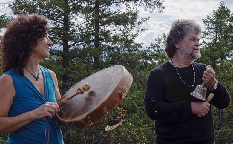 A white woman pounds a drum next to a white man holding a bell. They are standing in a forest.