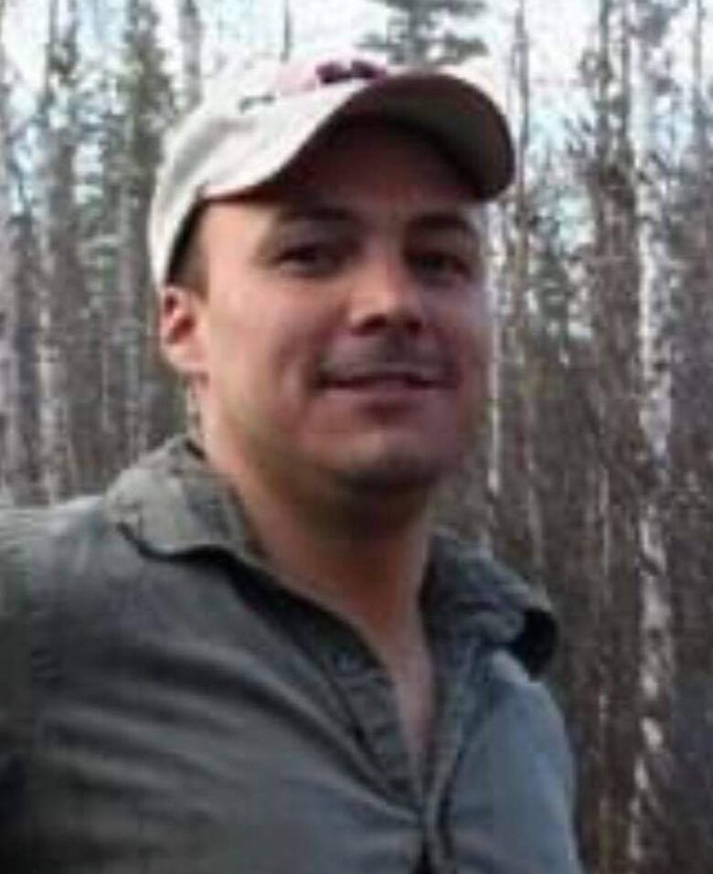 A man stands with a baseball cap. Trees in the background.