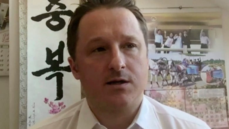 A man wearing a white collared shirt sits in front of a calendar.