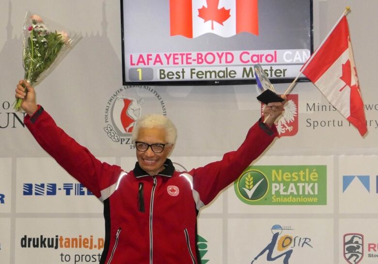 A woman stands with both arms raised, one holding a bouquet and the other a trophy and a Canadian flag.