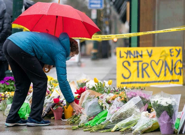 A person in black plants and a blue jacket holds a red umbrella as they place a bouquet of flowers on a pile of tributes on a concrete sidewalk. A yellow sign that says 