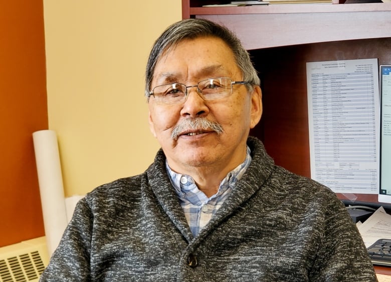 A man with grey hair and glasses, wearing a grey button-up sweater and a collared shirt underneath.