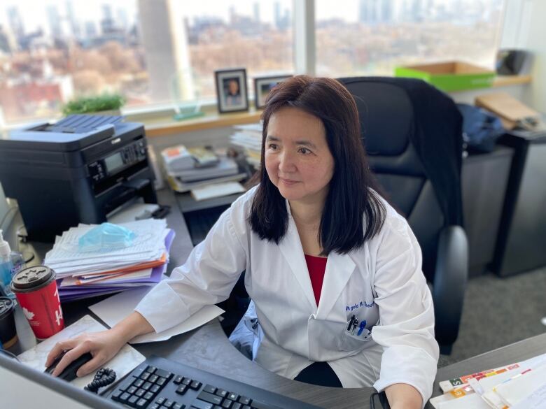 An Asian doctor wearing a lab coat works at a computer in her office. 