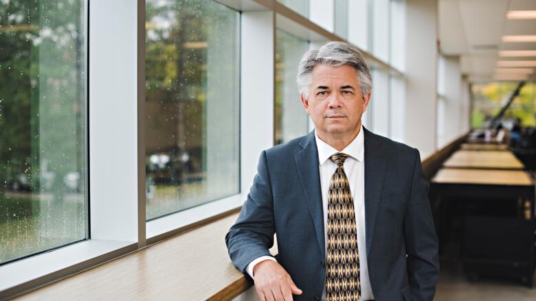 Man with grey hair wearing a suit and tie, standing inside a building with elbow resting on window sill.