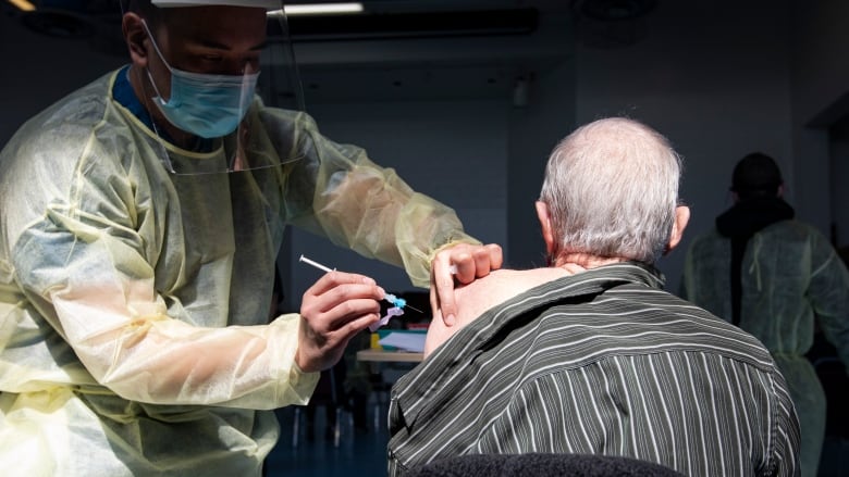 A health-care worker wearing personal protective equipment, including a face shield and mask, administers a vaccine into the arm of an elderly man.