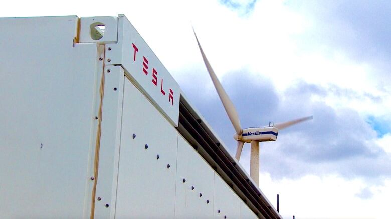 A Tesla-branded bank of batteries in the foreground, a giant wind turbine in the background.