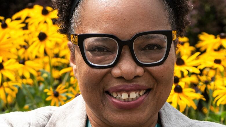 A Black woman is smiling outside in front of yellow flowers with brown centres.