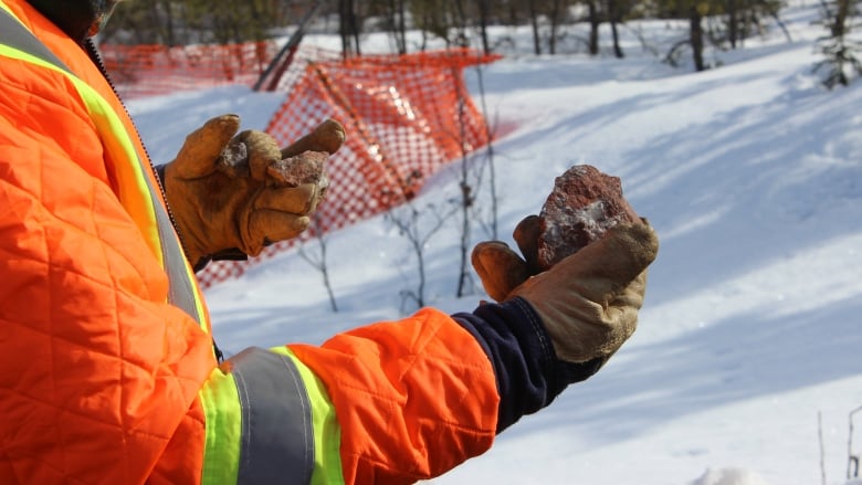 A close up of a person in a bright orange safety coat and gloves holding up a piece of rock.