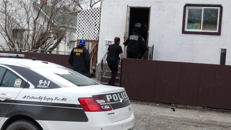 Police officers enter a white home, while a police vehicle is parked in front.