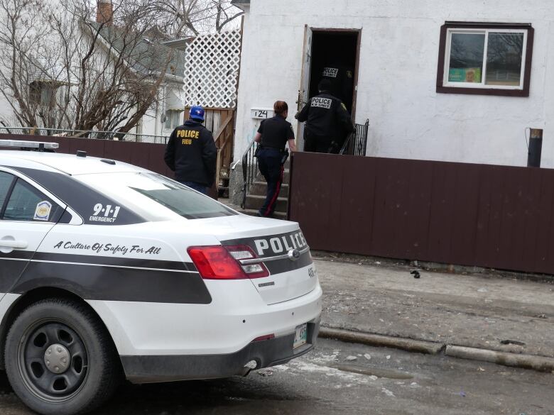 Police officers enter a white home, while a police vehicle is parked in front.