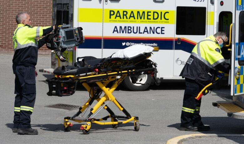 a Paramedic holds equipment beside a stretcher, while another grabs something from an open ambulance