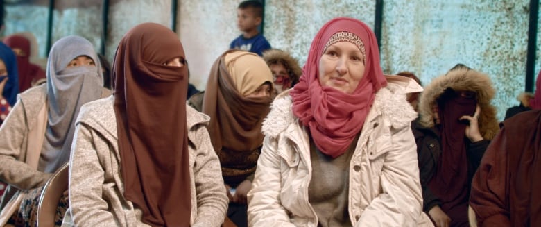 A white woman in a hijab speaks while sitting down. She is surrounded by woman wearing niqabs.