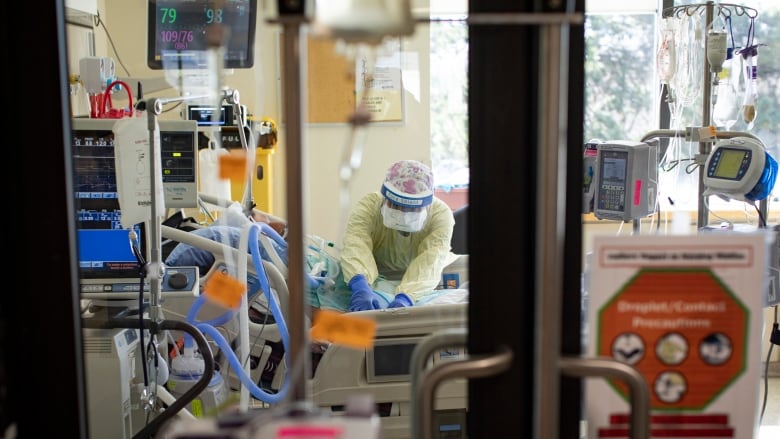 A nurse wearing personal protective equipment tends to a patient in the intensive care unit (ICU). They are masked as they are positioning a patient on a hospital bed surrounded by medical equipment. 