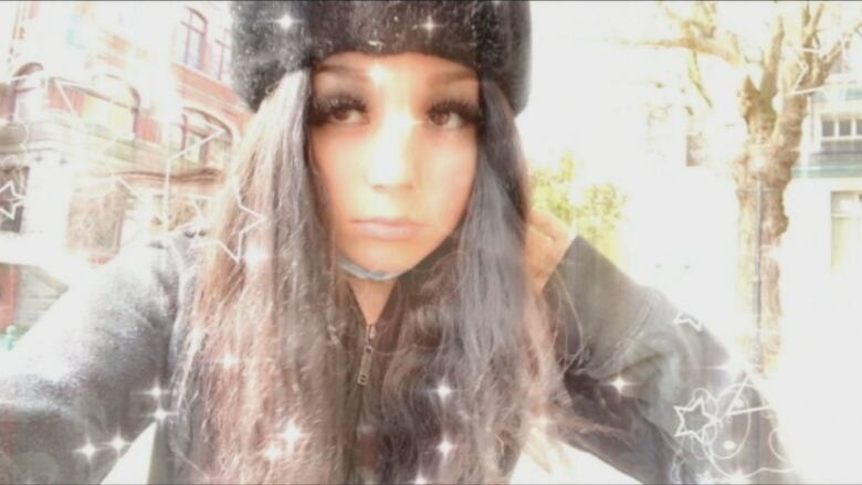 A young girl with long black hair, long and thick black eye lashes, a black hat and a dark jacket takes a selfie. A light filter of glimmers and outlines of stars is seen over the image and along its border.