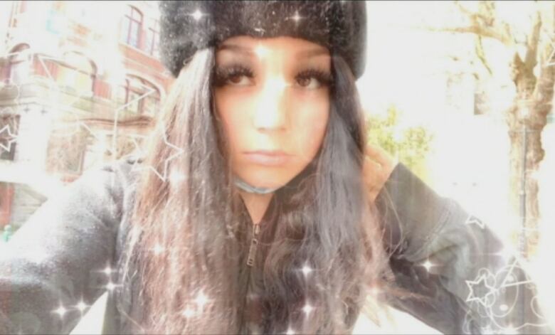 A young girl with long black hair, long and thick black eye lashes, a black hat and a dark jacket takes a selfie. A light filter of glimmers and outlines of stars is seen over the image and along its border.