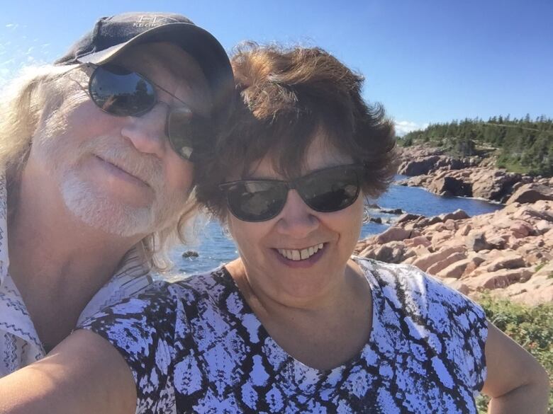 A man and woman take a selfie with a rocky ocean shoreline and blue sky in the background.