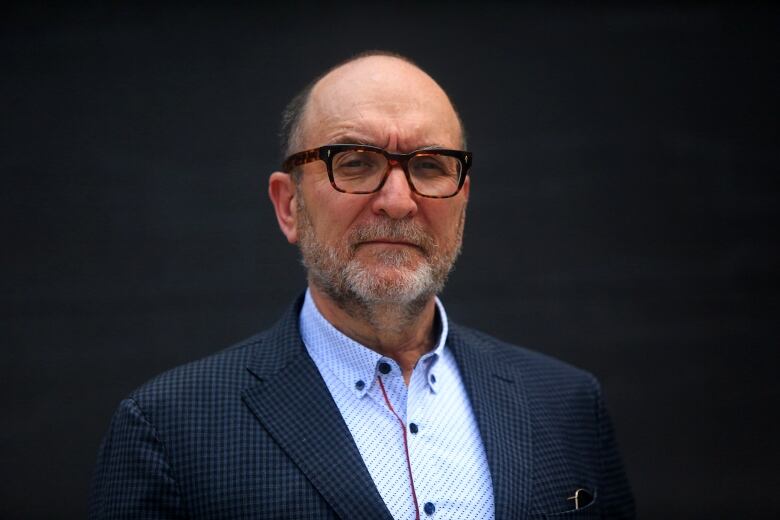 A man wearing glasses and a suit jacket and button-up shirt looks serious.