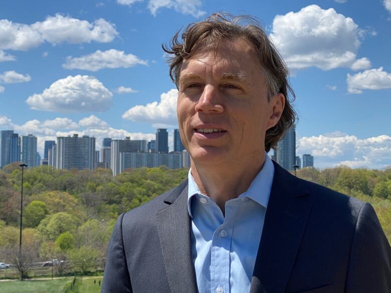 A man in a suit jacket and shirt stands in front of a skyline partially shrouded by trees