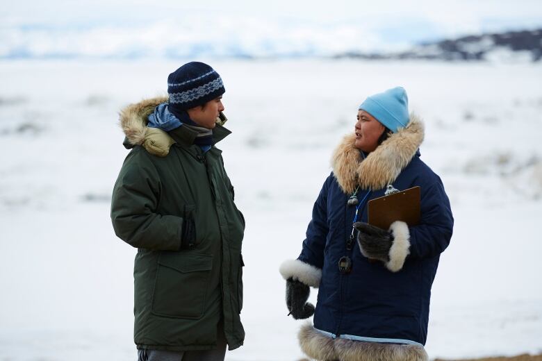 A man and a woman, wearing winter clothing stand and look at each other, the woman holding a clipboard in her hand 