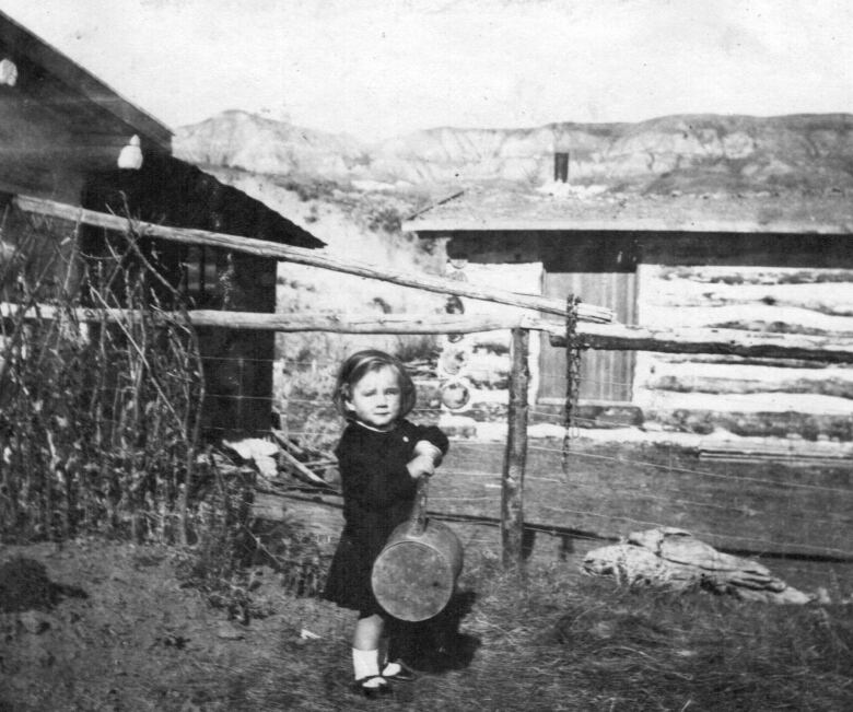 A young girl stands in a yard outside of log structures.