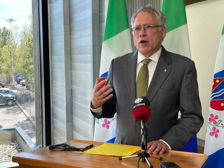 A man in a suit stands at a podium with a CBC microphone and a backdrop of Yukon flags.