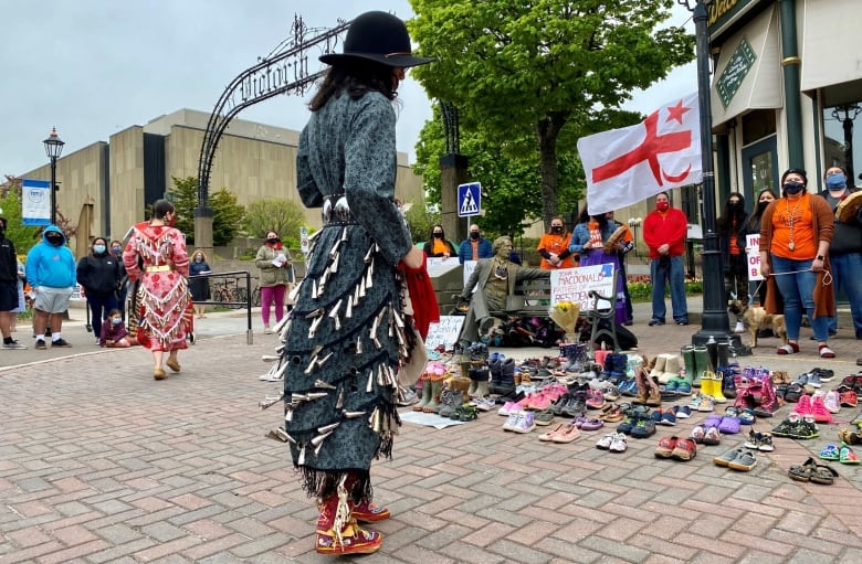 Jingle dancers dressed in regalia stand amongst a crowd of people surrounding the a seated statue of John A Macdonald. 