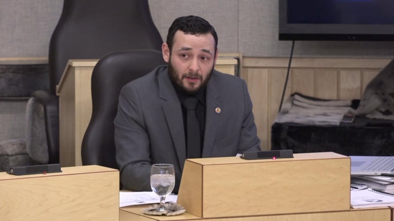 A man wearing a grey suit and black shirt and tie sitting in a legislative assembly