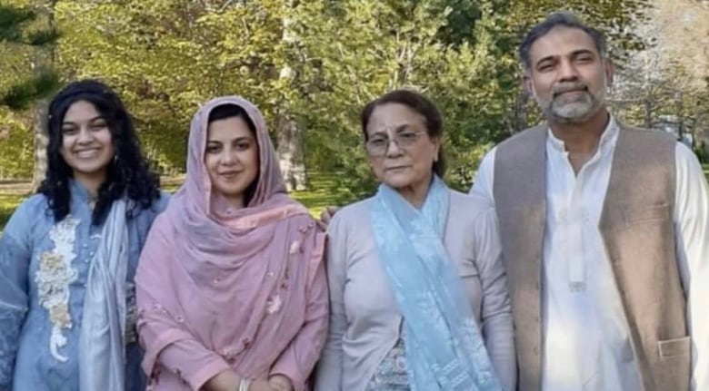 A family stands outside, looking to the camera. There are trees in the background.