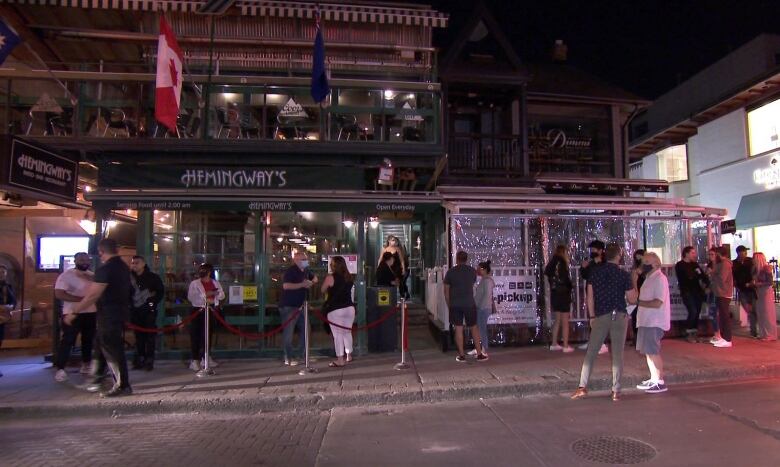 A crowd of people on a city street wait in line outside a bar.