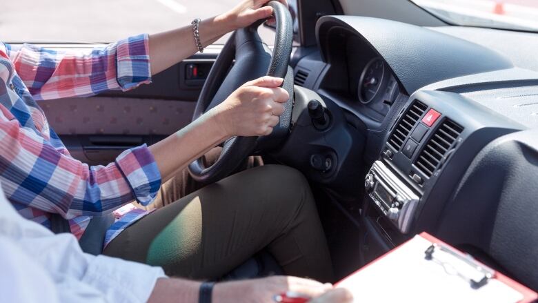 A driver wearing a plaid shirt sits at a steering wheel while an instructor holds a clipboard in the passenger seat. 