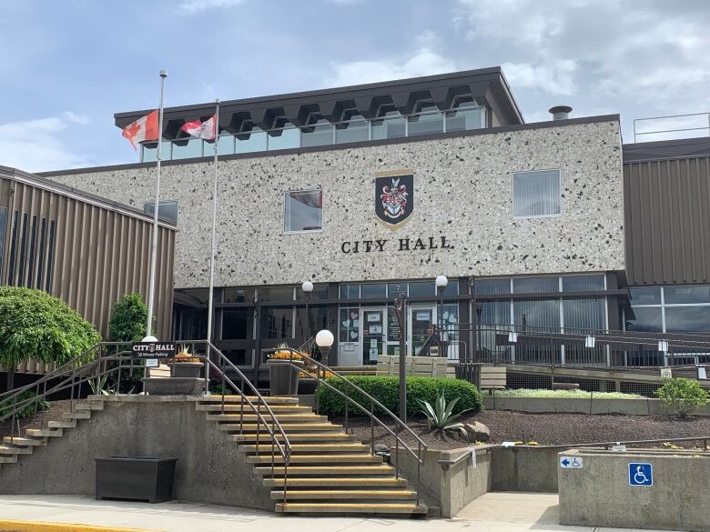 Brown stone steps lead up to a glass-fronted exterior below a white stone wall that says City Hall in black, topped by a coat of arms.