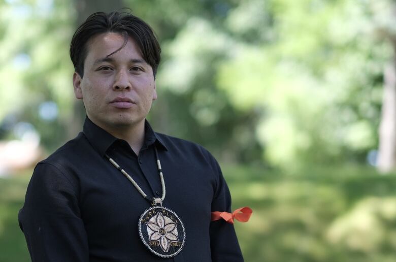 A man wearing a beaded medallion stands in a park.