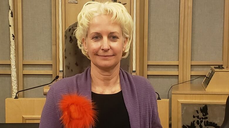 A smiling woman wearing a purple sweater stands in a territorial legislature.