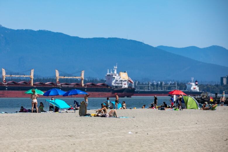 About 15 people sun tan and chat a beach on a hot sunny day, accompanied by blue beach umbrellas and towels.